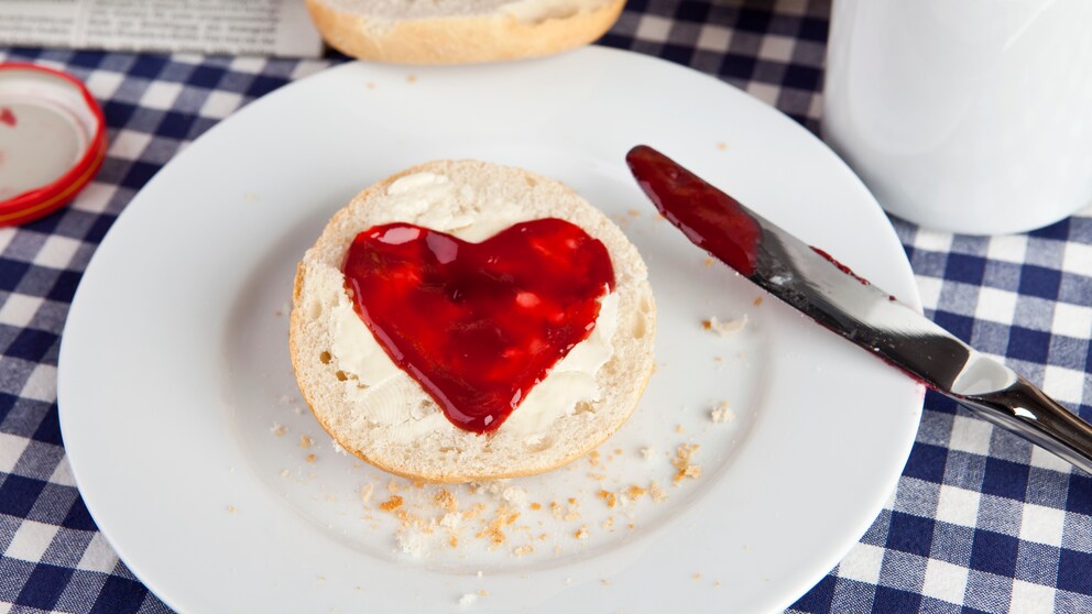 Auf einem Frühstückstisch liegt ein Brötchen mit einem Marmeladenaufstrich in Herzform