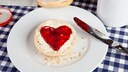 Auf einem Frühstückstisch liegt ein Brötchen mit einem Marmeladenaufstrich in Herzform