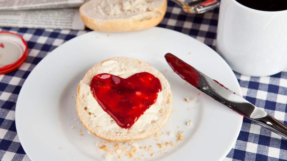 Auf einem Frühstückstisch liegt ein Brötchen mit einem Marmeladenaufstrich in Herzform