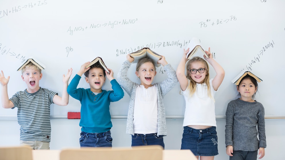 Fröhliche Mädchen und Jungen stehen vor einer Tafel, halten sich jeweils ein aufgeschlagenes Buch über den Kopf