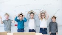 Fröhliche Mädchen und Jungen stehen vor einer Tafel, halten sich jeweils ein aufgeschlagenes Buch über den Kopf