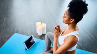 Ein junge Frau sitzt auf einer Sportmatte und meditiert, während vor ihr Kerzen und ein Tablet stehen