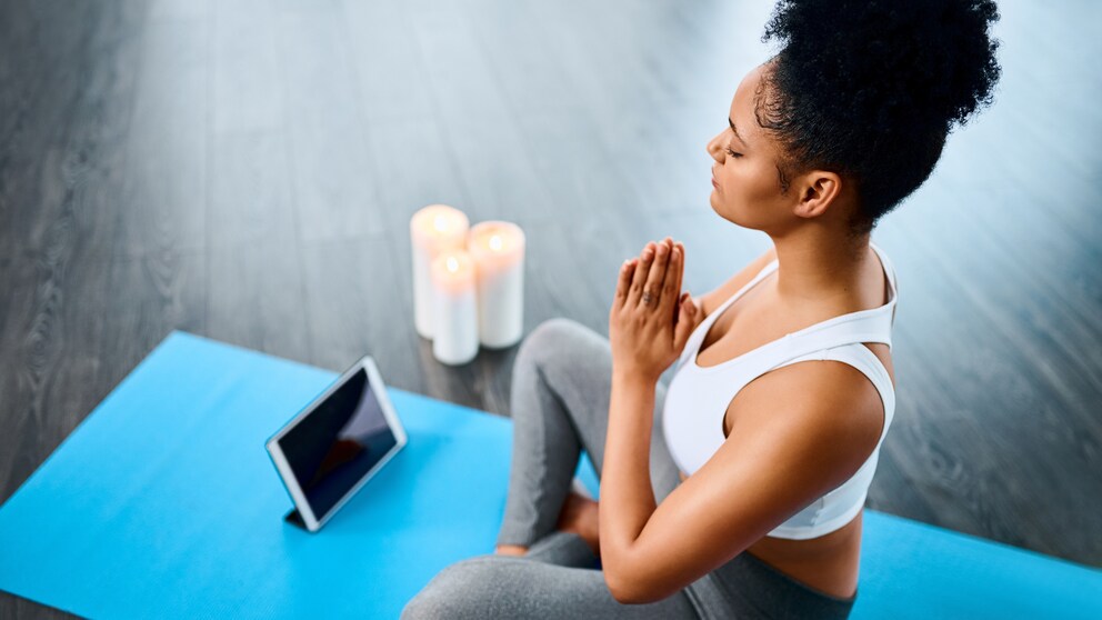 Ein junge Frau sitzt auf einer Sportmatte und meditiert, während vor ihr Kerzen und ein Tablet stehen