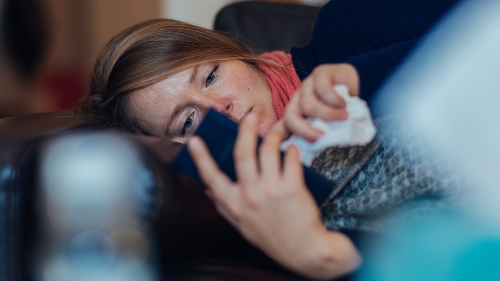 Corona-Verdacht: Eine kranke Frau liegt auf dem Sofa und hält ein Smartphone in der Hand