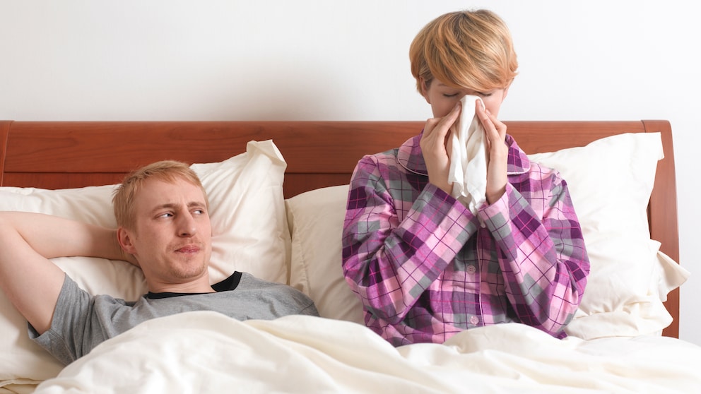 Niesen Sex: Ein Paar liegt im Bett, der Mann schaut die Frau an, während sie niest