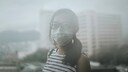 Luftverschmutzung Corona: Eine asiatische Frau tr&auml;gt eine Mund-Nasen-Schutzmaske, im Hintergrund Hochh&auml;user im Smog