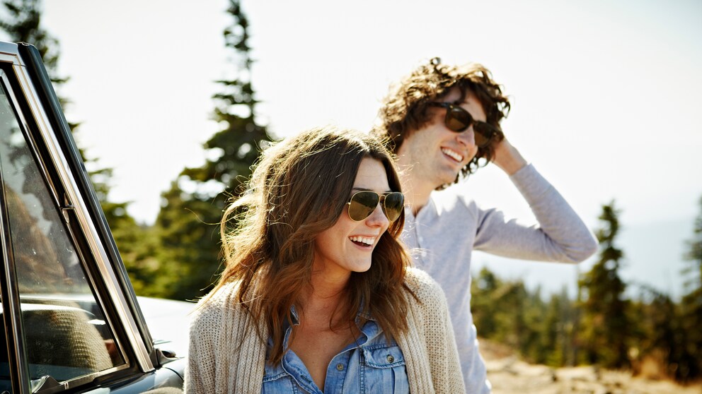 Ein junges Paar mit Sonnenbrille an Auto gelehnt in der Natur
