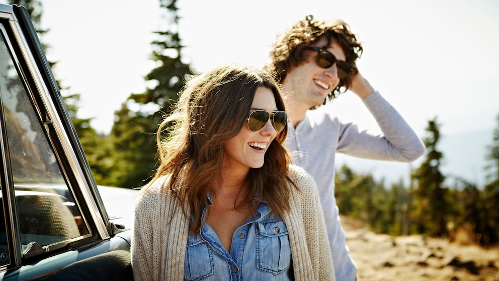Ein junges Paar mit Sonnenbrille an Auto gelehnt in der Natur
