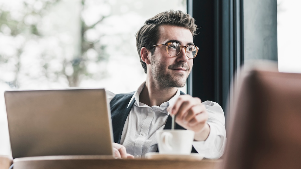 Junger Mann sitzt vor dem Laptop mit einer Tasse Kaffee