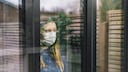 Junge Frau mit Mundschutz sieht aus dem Fenster