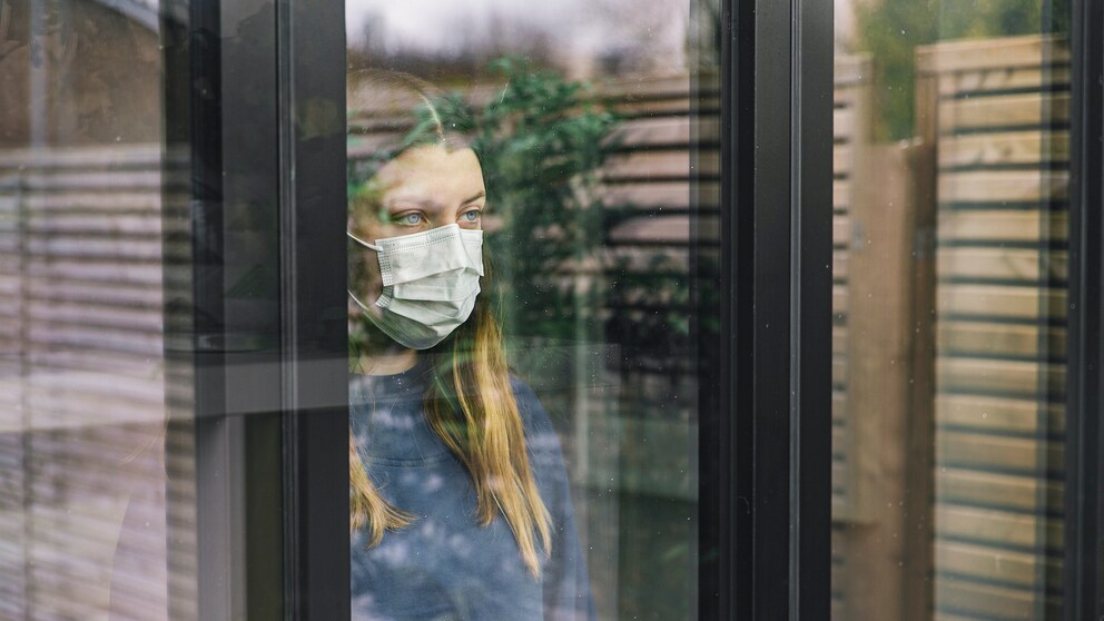 Junge Frau mit Mundschutz sieht aus dem Fenster