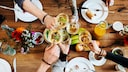 Vier Personen stoßen beim Essen am Tisch mit Wein an