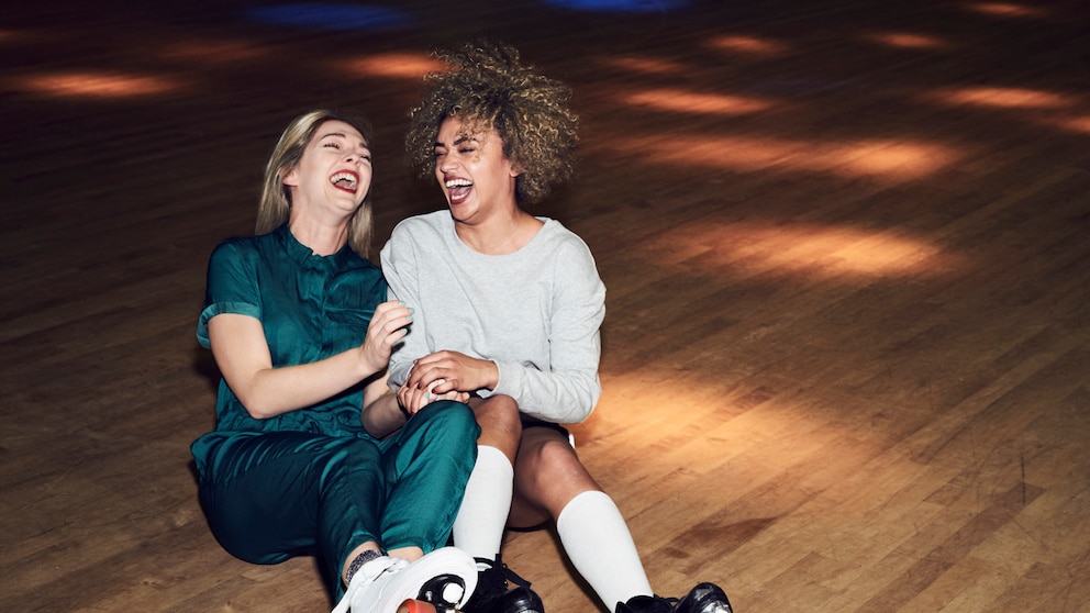 Zwei junge Frauen mit Rollschuhen sitzen lachend auf dem Boden