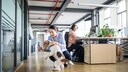 Eine Frau spielt im B&uuml;ro hockend mit einem Hund, w&auml;hrend ihre Kollegin an einem Schreibtisch sitzt