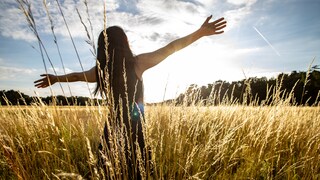 Frau steht mitten im Feld mir den Händen in der Luft und schaut gen Sonne.