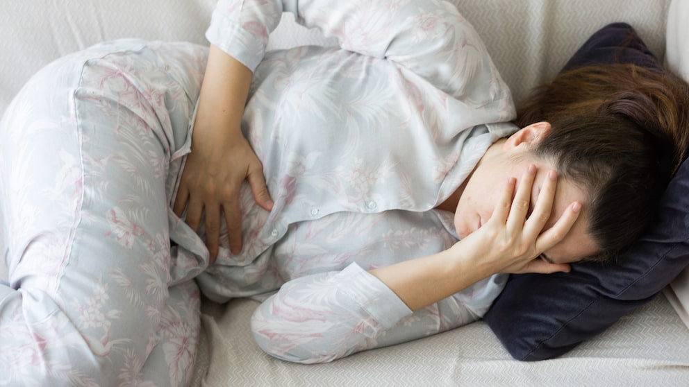 Frau liegt unter Schmerzen auf dem Sofa und hält sich den Bauch.