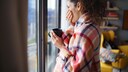 Junge Frau mit Decke umwickelt und tee in der Hand, steht am Fenster.