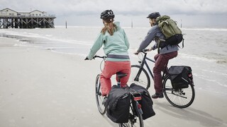 Zwei Personen mit Fahrradzubehör fahrend am Strand