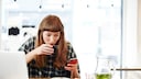 Junge Frage vor Laptop trinkt Kaffee und schaut aufs Smartphone