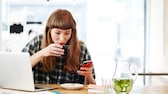 Junge Frage vor Laptop trinkt Kaffee und schaut aufs Smartphone