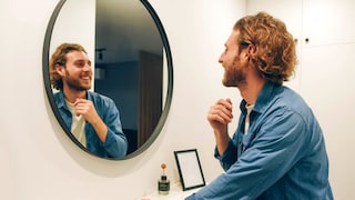 Handsome bearded guy looks at himself in the mirror. Jeans shirt. Brod guy. Admires himself. Nice appearance. Long hair. Casual style.