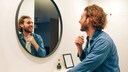 Handsome bearded guy looks at himself in the mirror. Jeans shirt. Brod guy. Admires himself. Nice appearance. Long hair. Casual style.