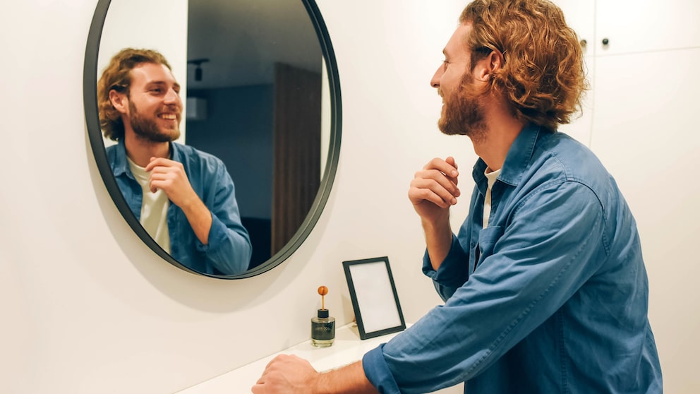 Handsome bearded guy looks at himself in the mirror. Jeans shirt. Brod guy. Admires himself. Nice appearance. Long hair. Casual style.