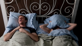 Pille gegen Schlafapnoe? Ein Mann liegt schnarchend neben einer Frau im Bett, die sich ein Kissen über den Kopf gelegt hat