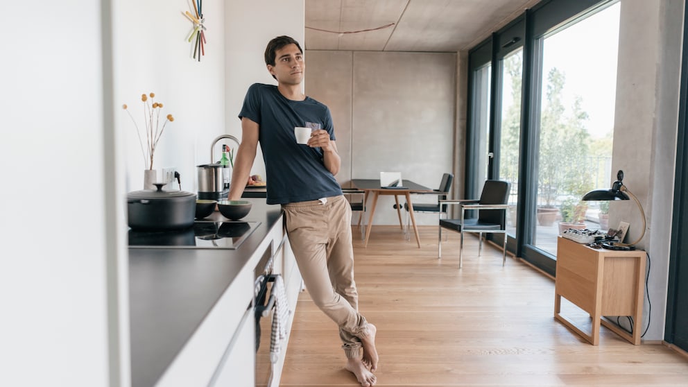 Ein Mann lehnt an einer Küchenzeile, hält einen Becher Kaffee in der Hand und sieht gedankenverloren aus dem Fenster