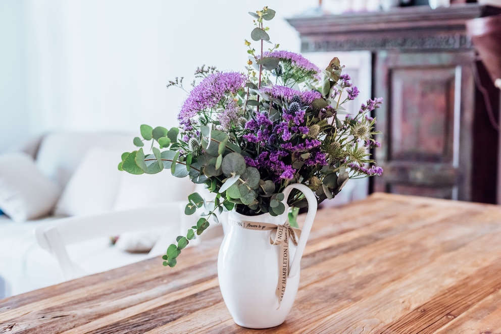 Auf einem Tisch steht eine Vase mit lilafarbenen Blumen und Gräsern