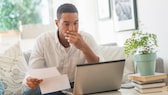 Mann sitzt auf dem Sofa mit einem Schreiben in der Hand und schaut erschrocken mit der linken Hand vor dem Mund auf den aufgeklappten Laptop vor ihm