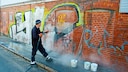 Ein Mann reinigt eine mit Graffiti bespr&uuml;hte rote Backsteinwand mit einem Hochdruckreiniger.