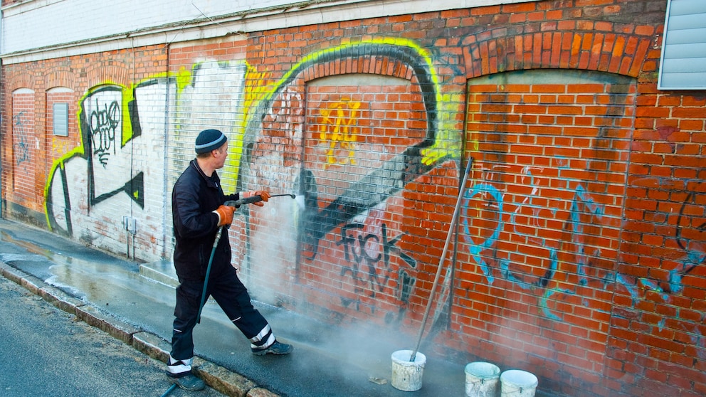 Ein Mann reinigt eine mit Graffiti besprühte rote Backsteinwand mit einem Hochdruckreiniger.