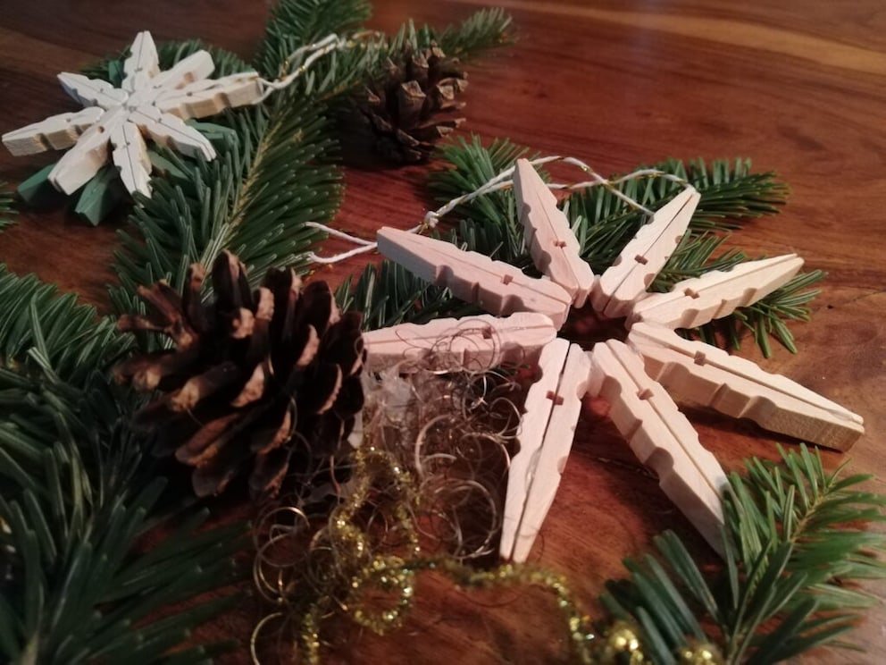 Christmas stars made from clothespins lie on a table between pine greenery and pine cones