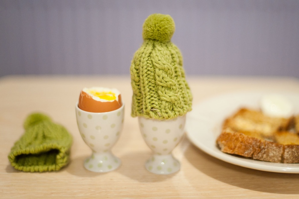 Grüne Eierwärmer in Form einer Pudelmütze neben einem geköpften Ei und einem Teller mit einem belegten Brot