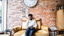Ein Mann sitzt auf einer Couch, über ihm eine Wanduhr