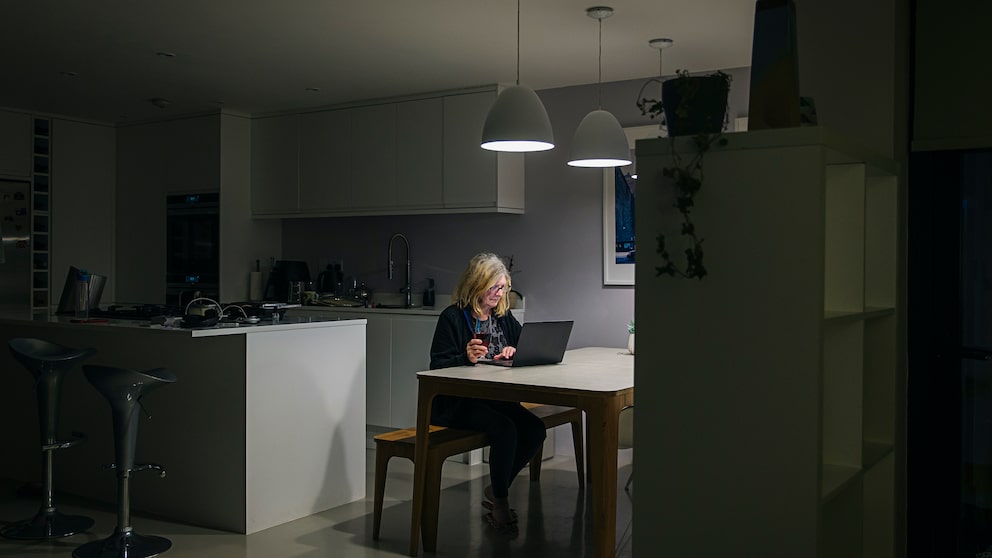 Frau, die in einer dunklen, innenliegenden Küche ohne Fenster, sitzt und am Laptop arbeitet