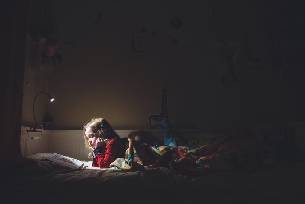 A child lying in bed in the dark, reading a book with a reading light