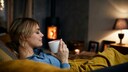 Frau sitzt auf einem Sofa vor dem Kamin, mit einer Tasse in der Hand und gehüllt in eine Decke
