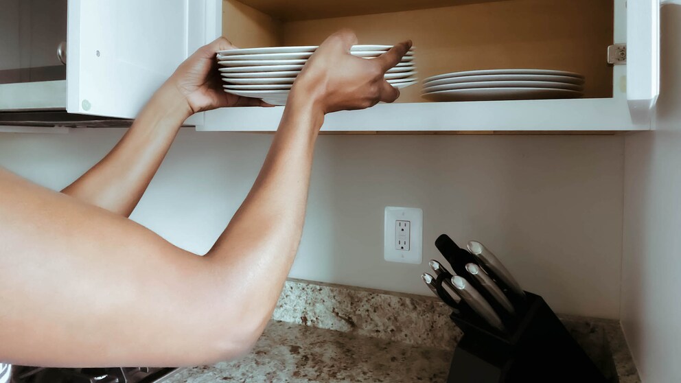 Nahaufnahme einer nicht erkennbaren Frau, die sauberes Geschirr auf ein Regal im Küchenschrank stellt