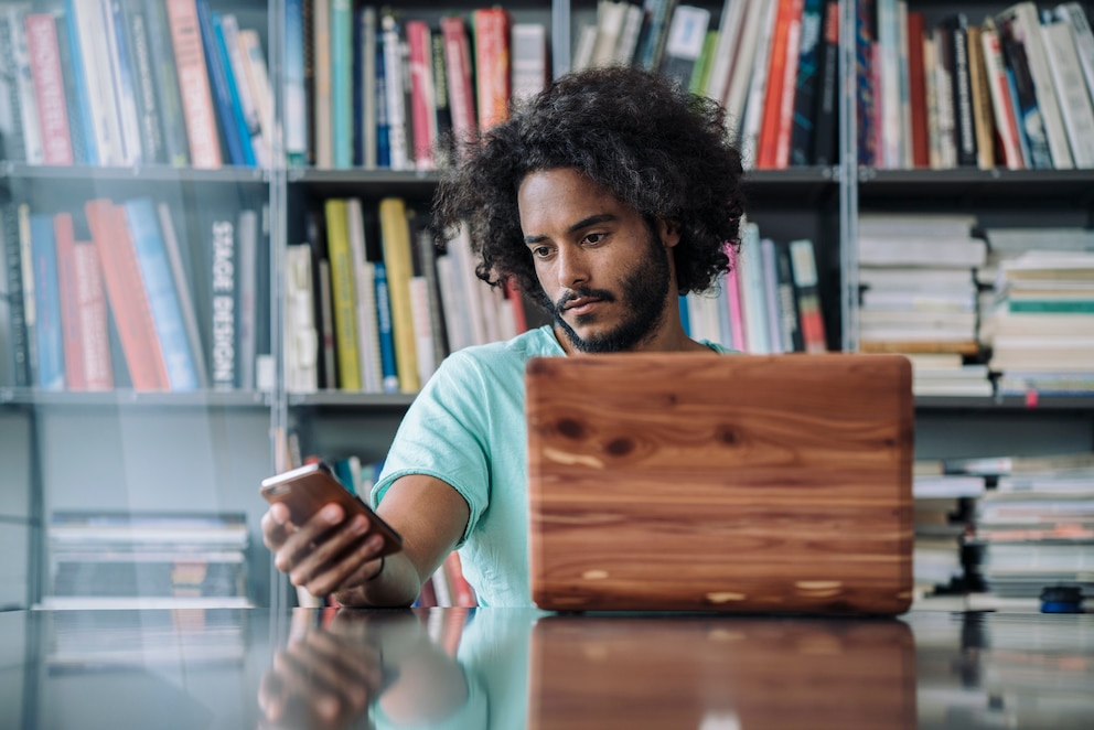 Junger Mann, der mit Smartphone und Laptop vor einem vollen Bücherregal sitzt