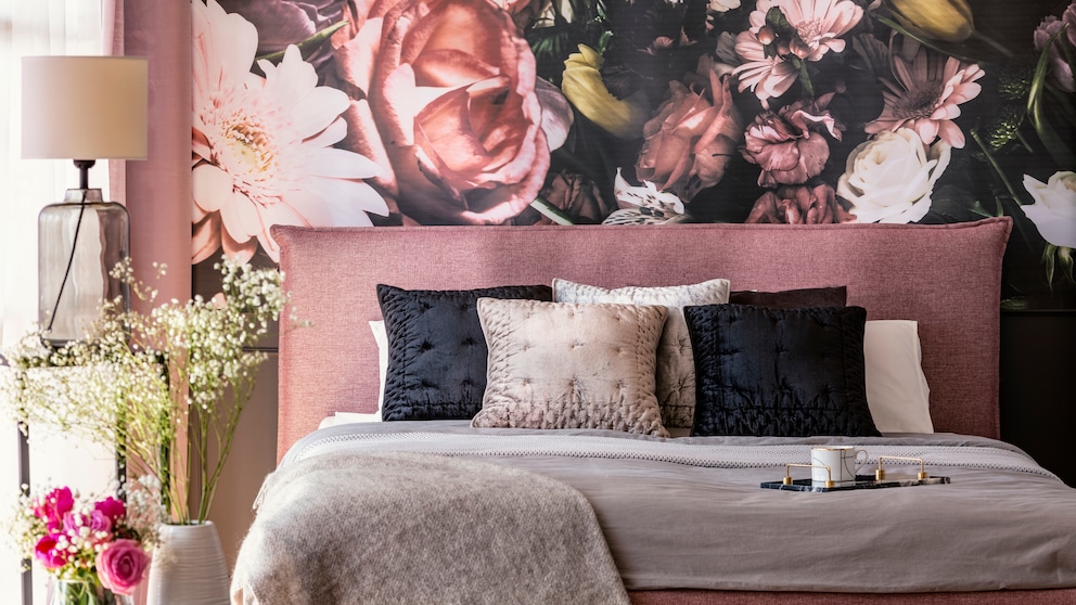 Ein textilreiches bett in Altrosa vor einer Tapete mit üppigem Blumenbouquet