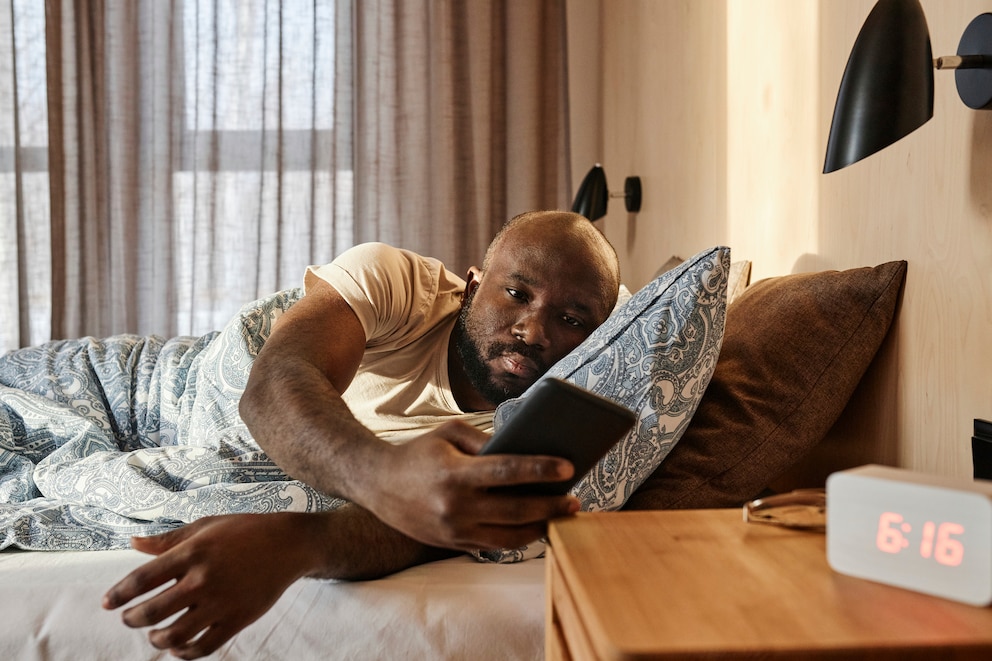 Mann, der im Bett liegt, schaut auf sein Smartphone, das auf dem Nachttisch neben dem Wecker liegt