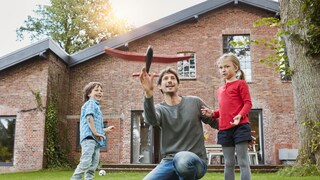 Ein Vater, der mIt seinen Kindern vor dem eigenen haus spielt. Für viele Deutsche platzt der Traum vom, Eigenheim, aufgrund der finanziellen Situation