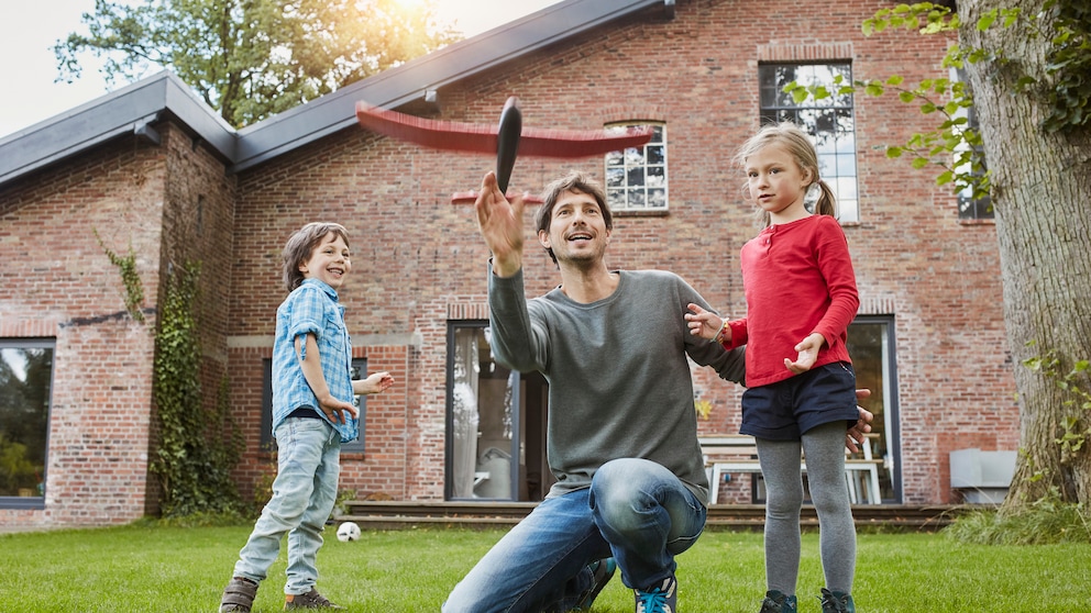 Ein Vater, der mIt seinen Kindern vor dem eigenen haus spielt. Für viele Deutsche platzt der Traum vom, Eigenheim, aufgrund der finanziellen Situation