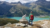 Ein Hund und seine Halterin in Wanderausrüstung stehen an einem klaren Bergsee