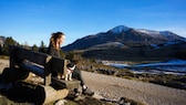 Reise-Autorin Sophie Lübbert auf einer Bank mit Hund Sam blickt auf Naturkulisse mit Fluss und Bergen