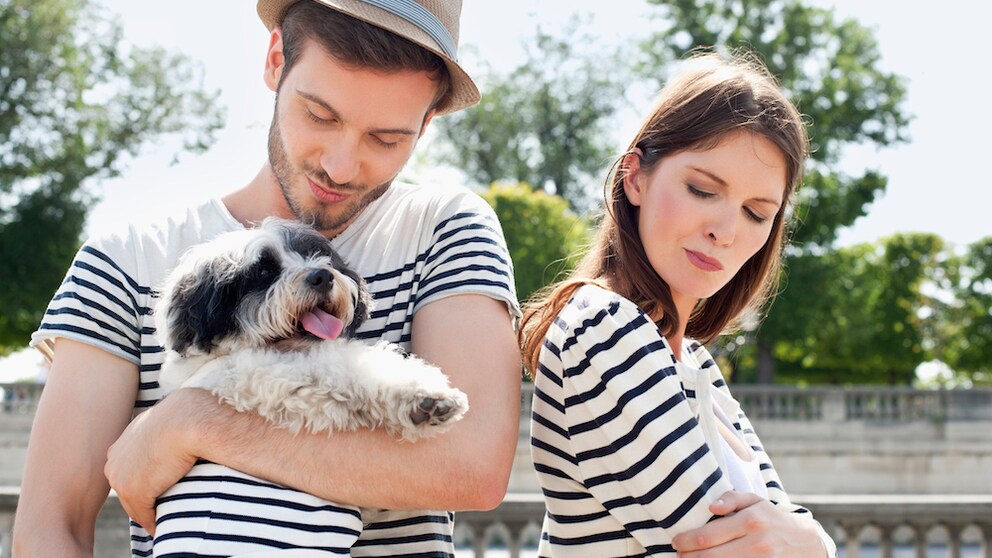 Man hat Hund auf dem Arm, Frau daneben schaut missbilligend zu den beiden