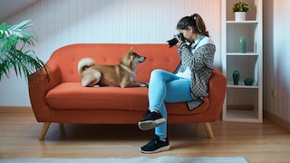 Frau hält eine Kamera in der Hand und sitzt Hund auf einem roten Sofa.