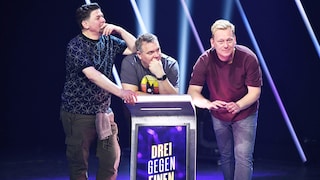 Tim Mälzer (l-r), Elton und Jens „Knossi“ Knossalla stehen an einem Podest im Studio bei der Show „Drei gegen Einen - Die Show der Champions“